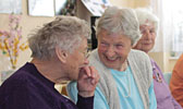 Foto: Zwei Seniorinnen sitzen am Tisch, essen Kuchen, lachen und unterhalten sich. Dieses Foto ist gleichzeitig ein Link und führt Sie zu der Unterseite: Seniorengruppe.